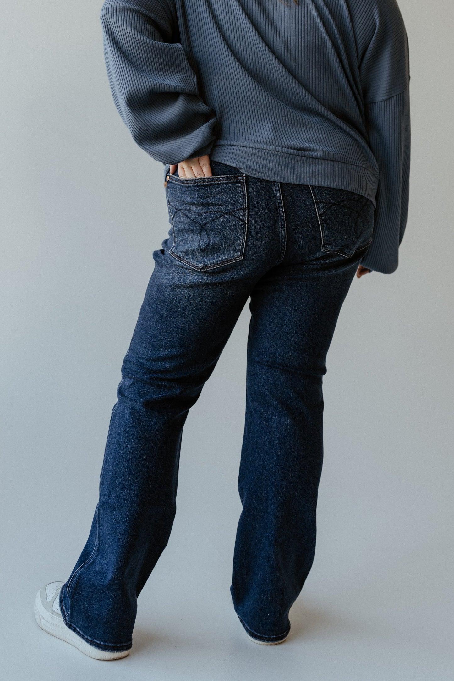 A person in a dark sweater and blue jeans with an embroidered pocket stands with one hand in their back pocket against a plain background.