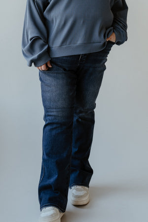 A person wearing a blue sweater, bootcut jeans, and white sneakers stands with hands in embroidered pockets against a plain background.