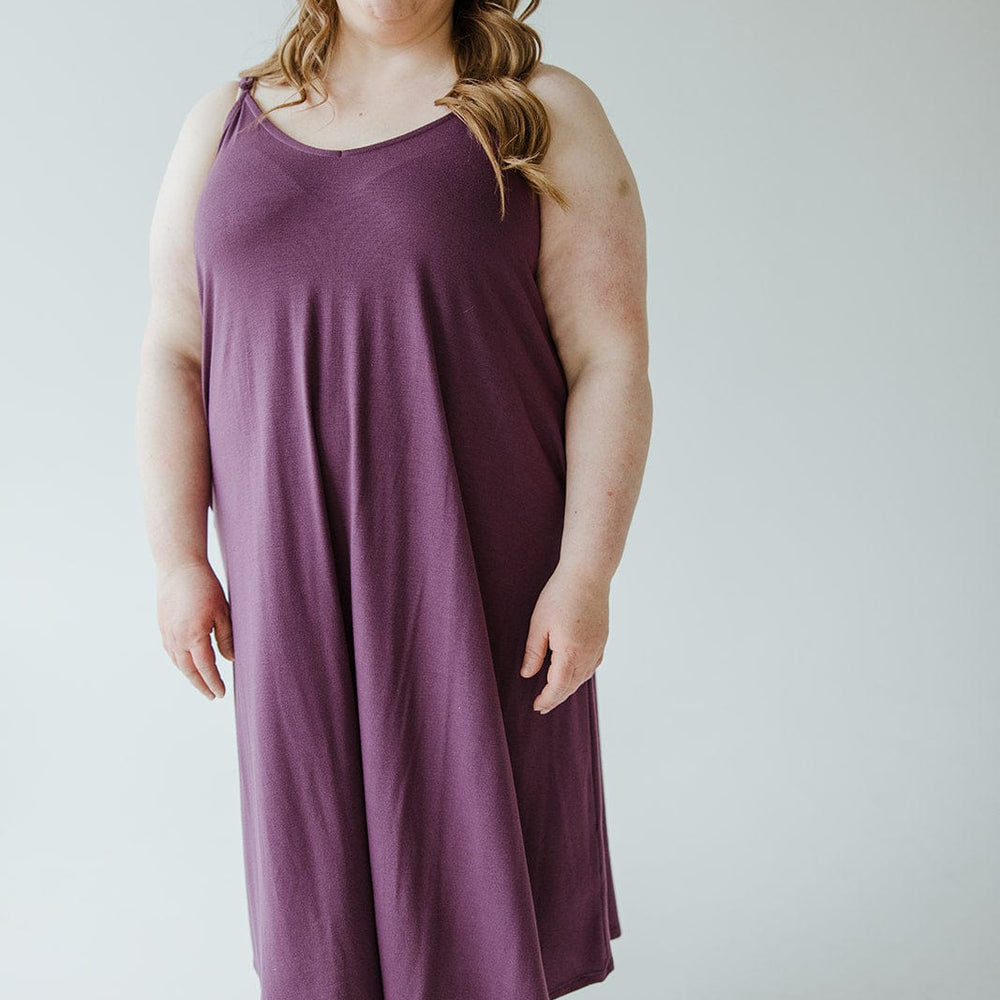 A person with long hair wearing an A-line cami dress in a rich antique violet hue stands against a plain background. The ultra-soft knit fabric adds a touch of elegance to their stylish look.