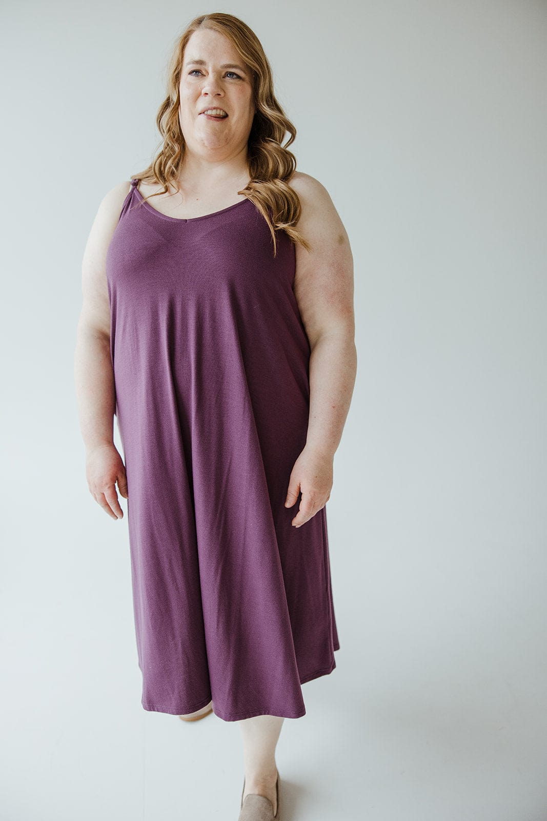 A person with long hair wearing an A-line cami dress in a rich antique violet hue stands against a plain background. The ultra-soft knit fabric adds a touch of elegance to their stylish look.