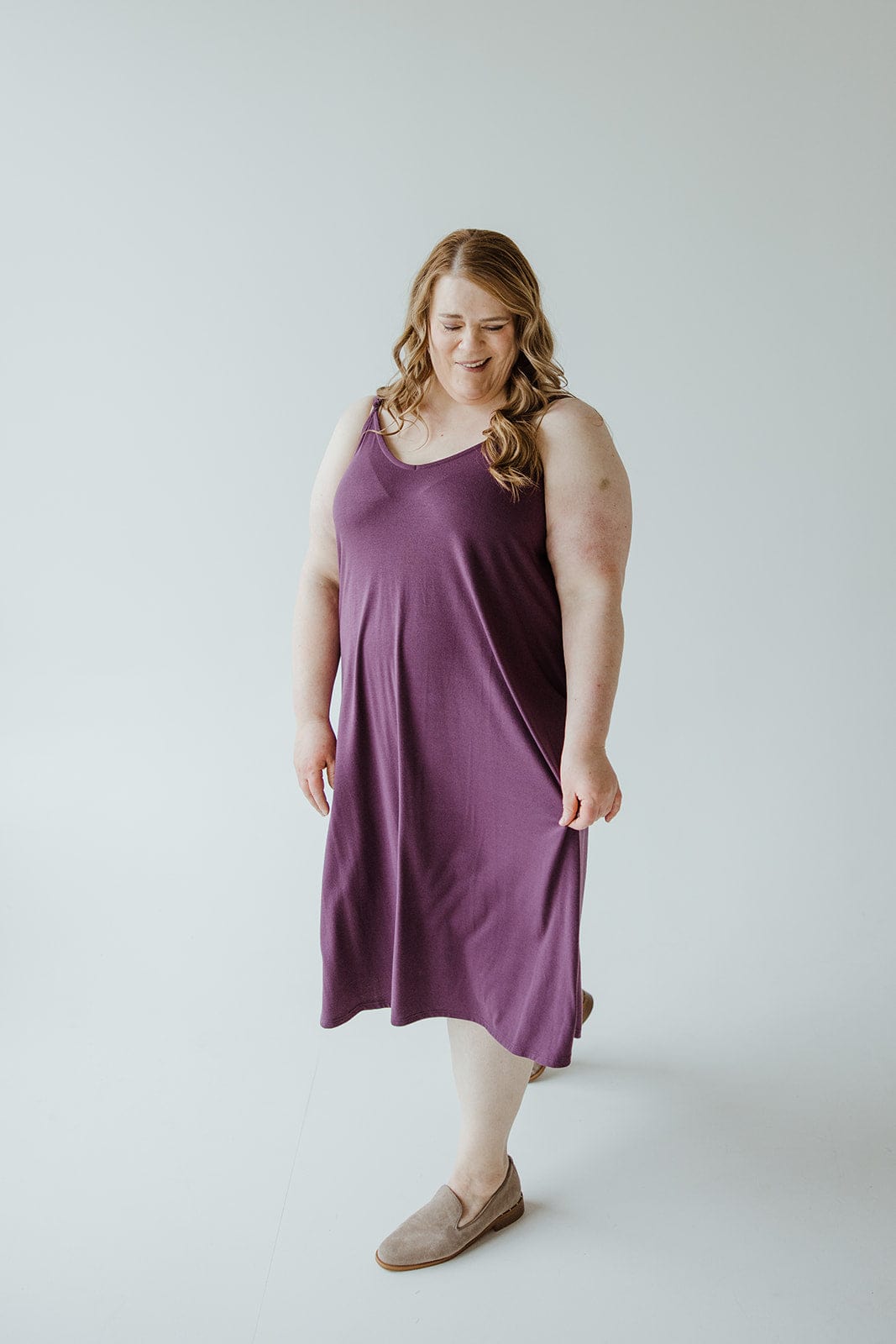 A person in an A-line cami dress of antique violet hue and brown slip-on shoes stands against a plain white background.