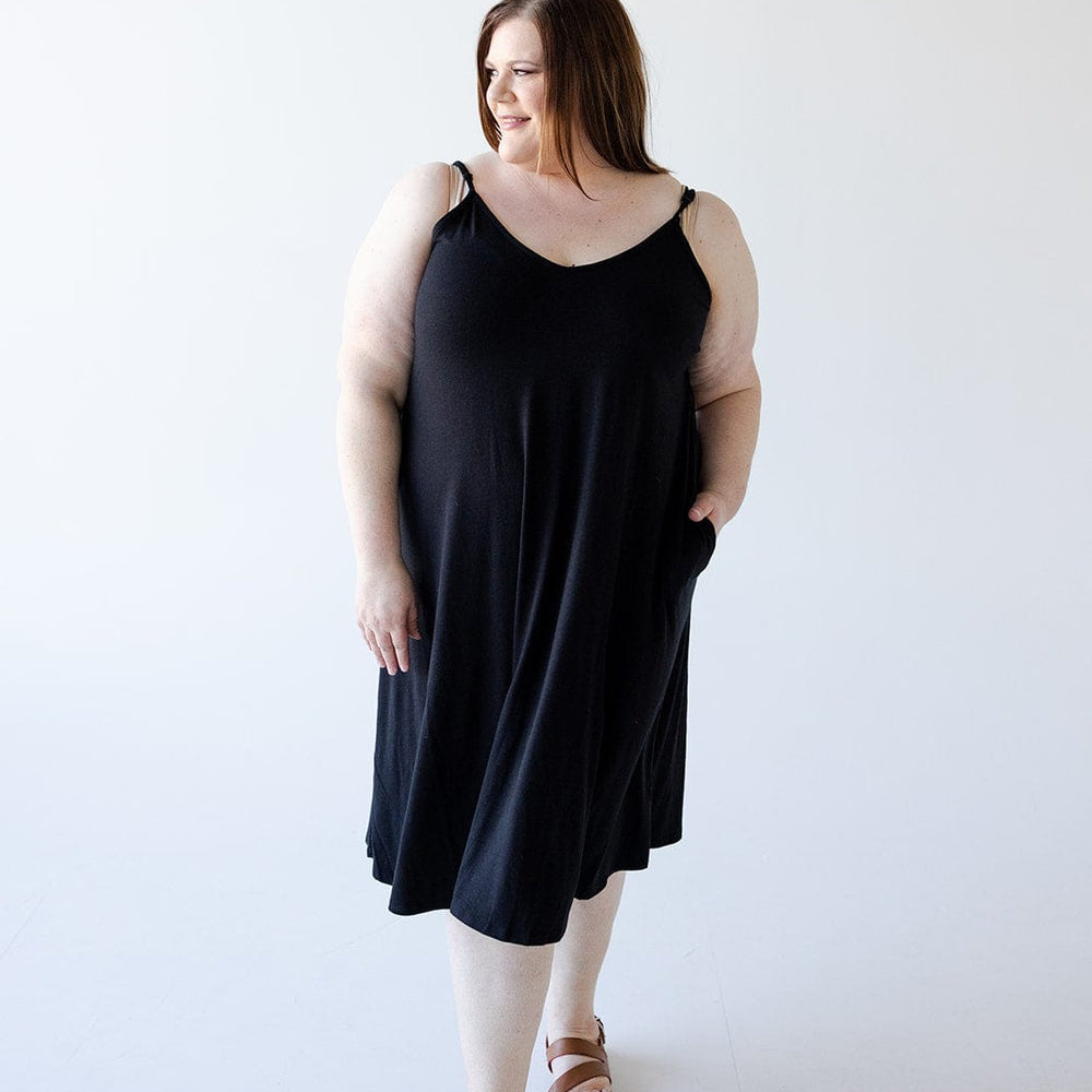 A woman stands posing in a figure-flattering Zenana Knee Length A-Line Cami Dress in Black, which features adjustable straps. She is looking slightly to her right and has one hand tucked into her pocket, complementing the outfit with brown sandals. The background is plain and white, emphasizing the simplicity of the dress.