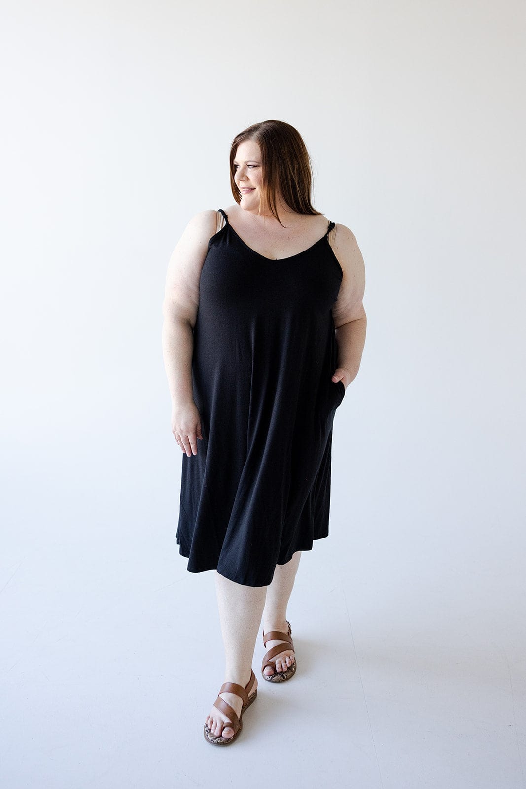 A woman stands posing in a figure-flattering Zenana Knee Length A-Line Cami Dress in Black, which features adjustable straps. She is looking slightly to her right and has one hand tucked into her pocket, complementing the outfit with brown sandals. The background is plain and white, emphasizing the simplicity of the dress.