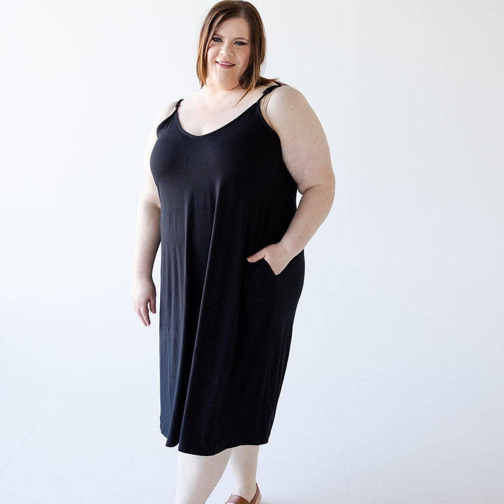 A woman stands against a plain white background, wearing the Zenana Knee Length A-Line Cami Dress in Black with adjustable straps, beige leggings, and brown sandals. She has one hand in the pocket of her figure-flattering dress and smiles at the camera.