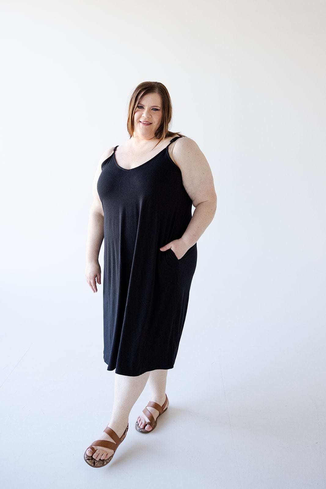 A woman stands against a plain white background, wearing the Zenana Knee Length A-Line Cami Dress in Black with adjustable straps, beige leggings, and brown sandals. She has one hand in the pocket of her figure-flattering dress and smiles at the camera.