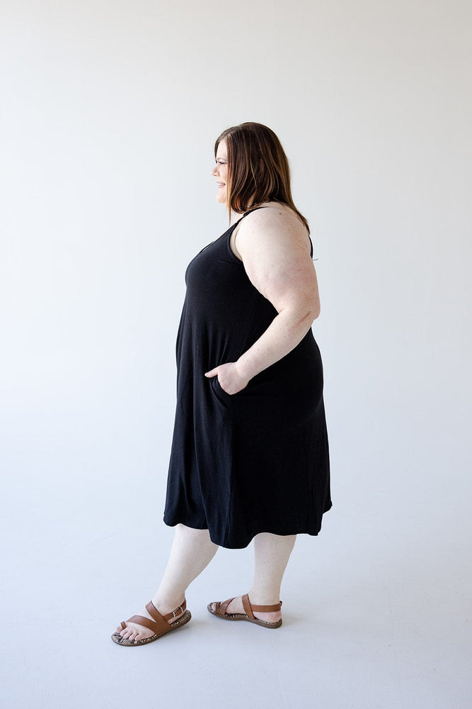 A woman stands in profile, wearing the Zenana Knee Length A-Line Cami Dress in Black with adjustable straps and sandals. Her hand is casually placed in the dress pocket, set against a plain white background.