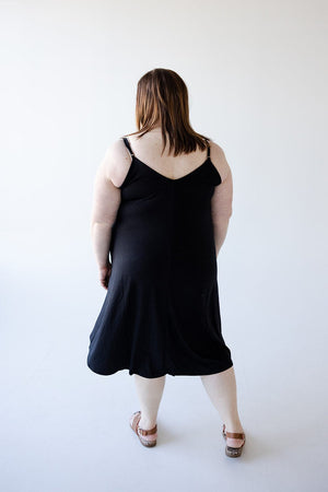 A person with shoulder-length hair, wearing Zenana's figure-flattering KNEE LENGTH A-LINE CAMI DRESS IN BLACK with adjustable straps and sandals, is standing with their back toward the camera against a plain white background.