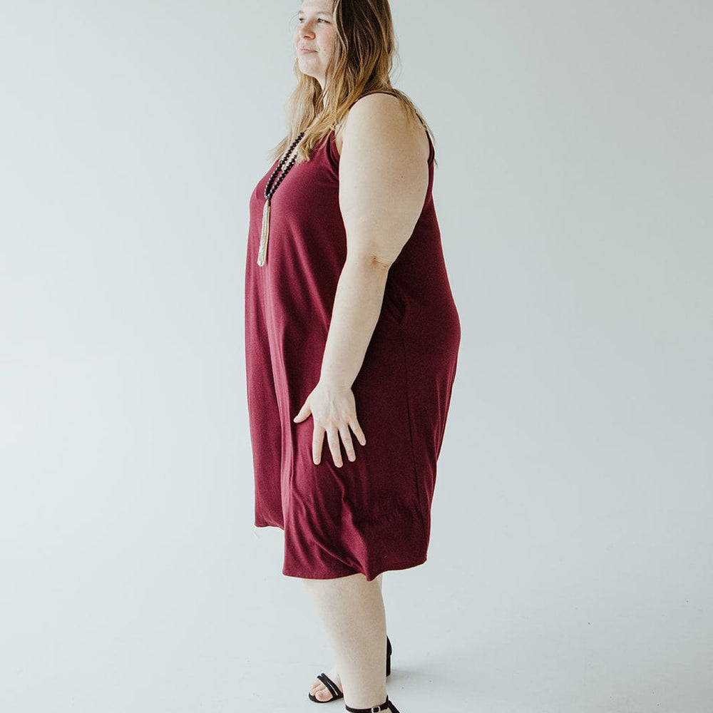 A person in a dark red cami dress with adjustable straps and black sandals stands against a plain light background, looking to the left.