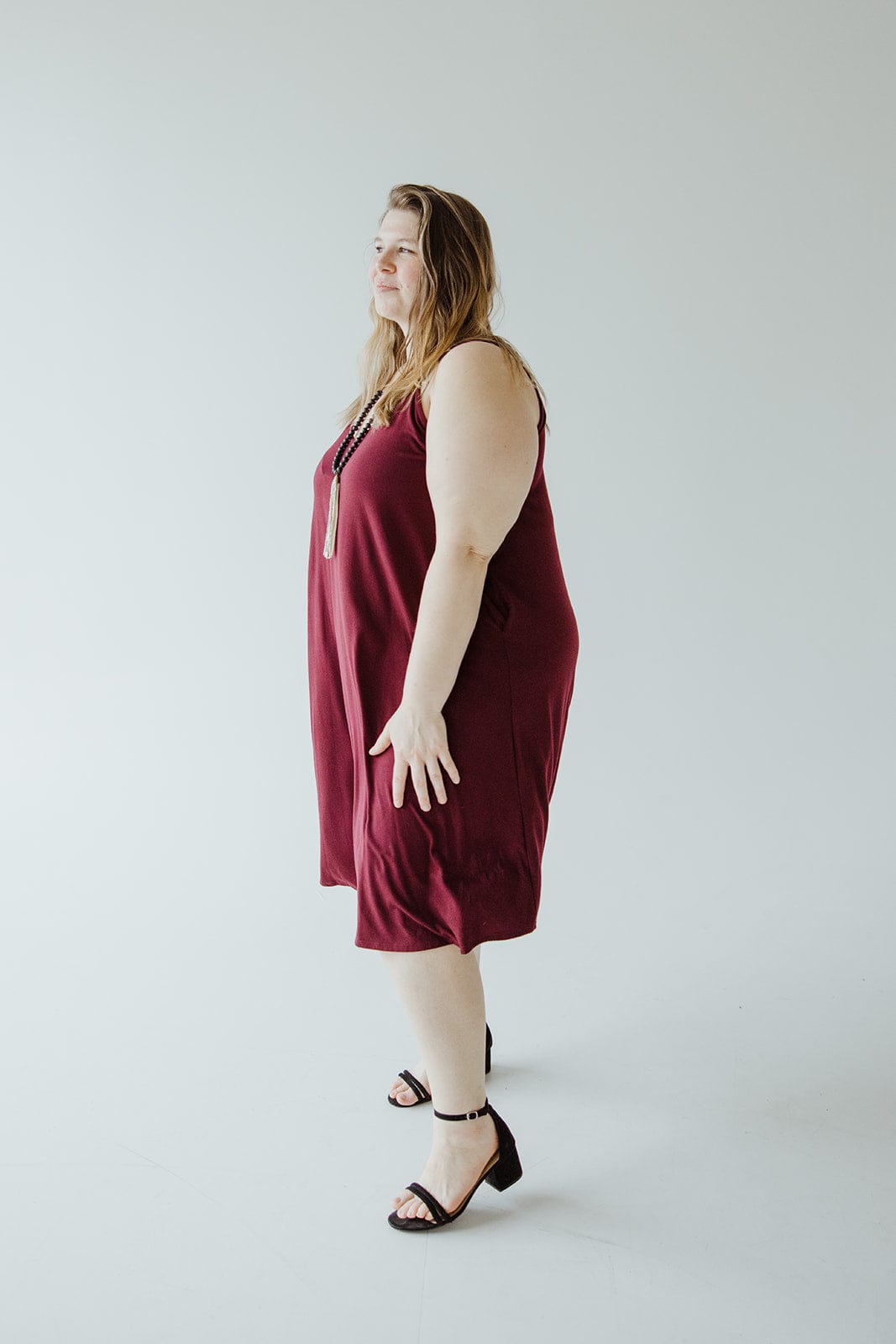 A person in a dark red cami dress with adjustable straps and black sandals stands against a plain light background, looking to the left.