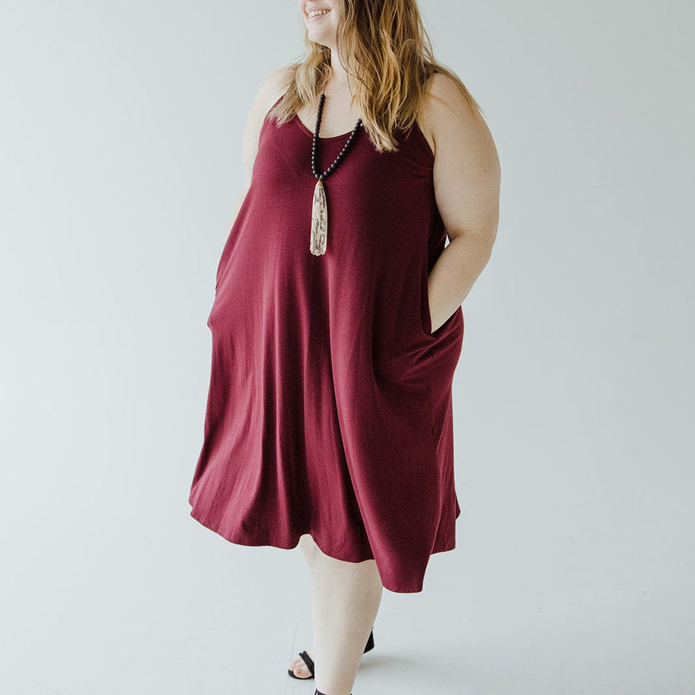 A person in a sleeveless burgundy cami dress featuring v-neck detail stands with hands in pockets, wearing black sandals and a long necklace.