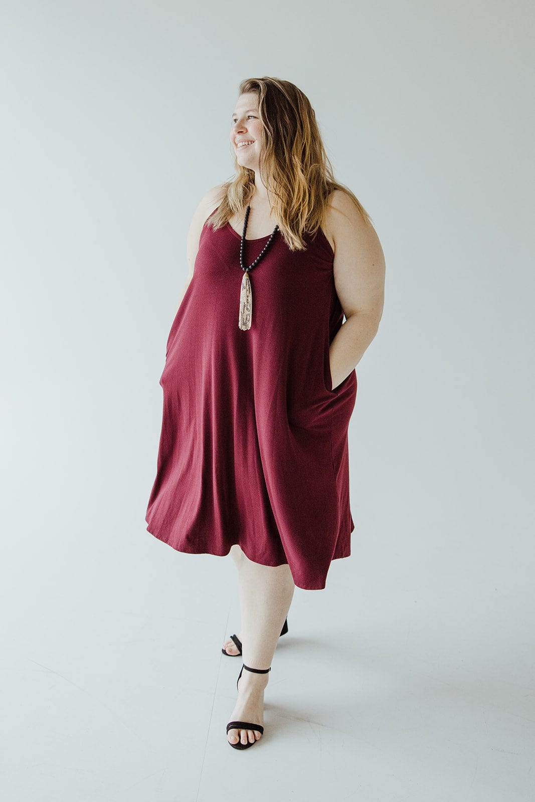 A person in a sleeveless burgundy cami dress featuring v-neck detail stands with hands in pockets, wearing black sandals and a long necklace.