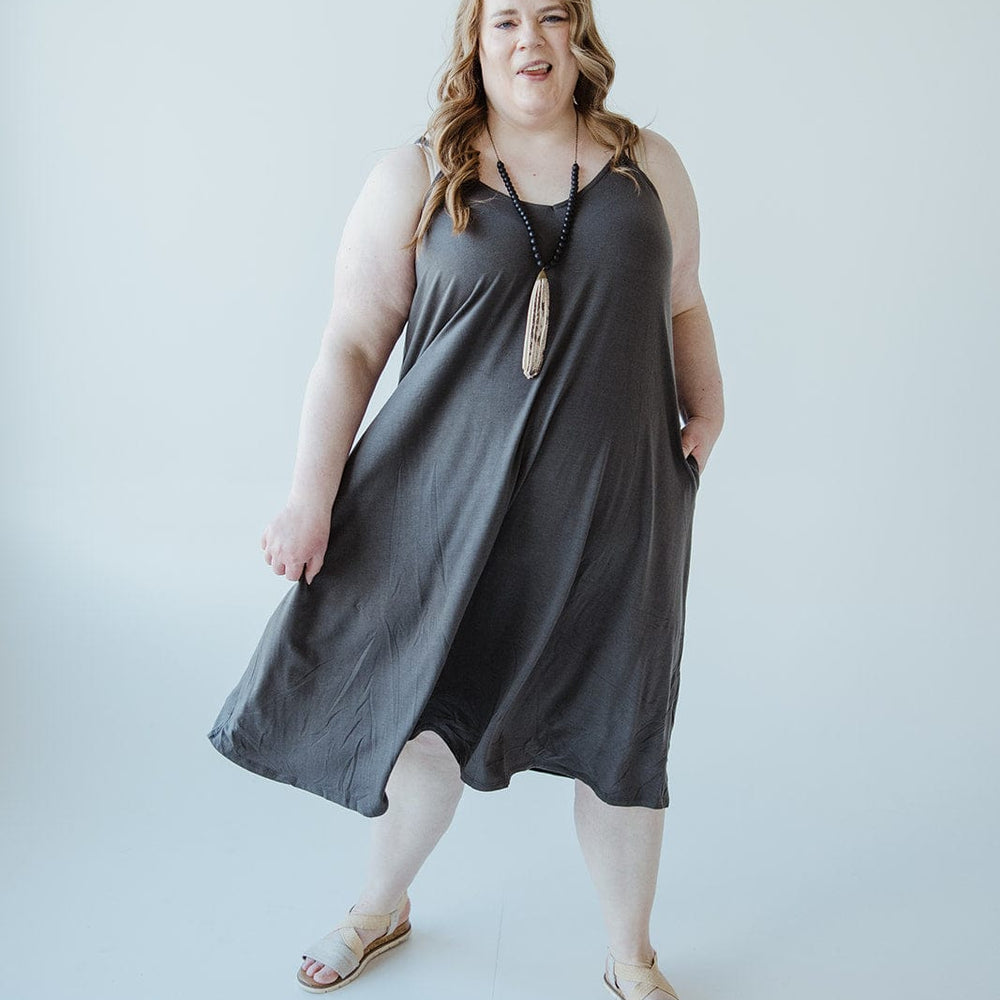 A person with long hair stands against a plain background, wearing a charcoal-hued cami dress paired with sandals.