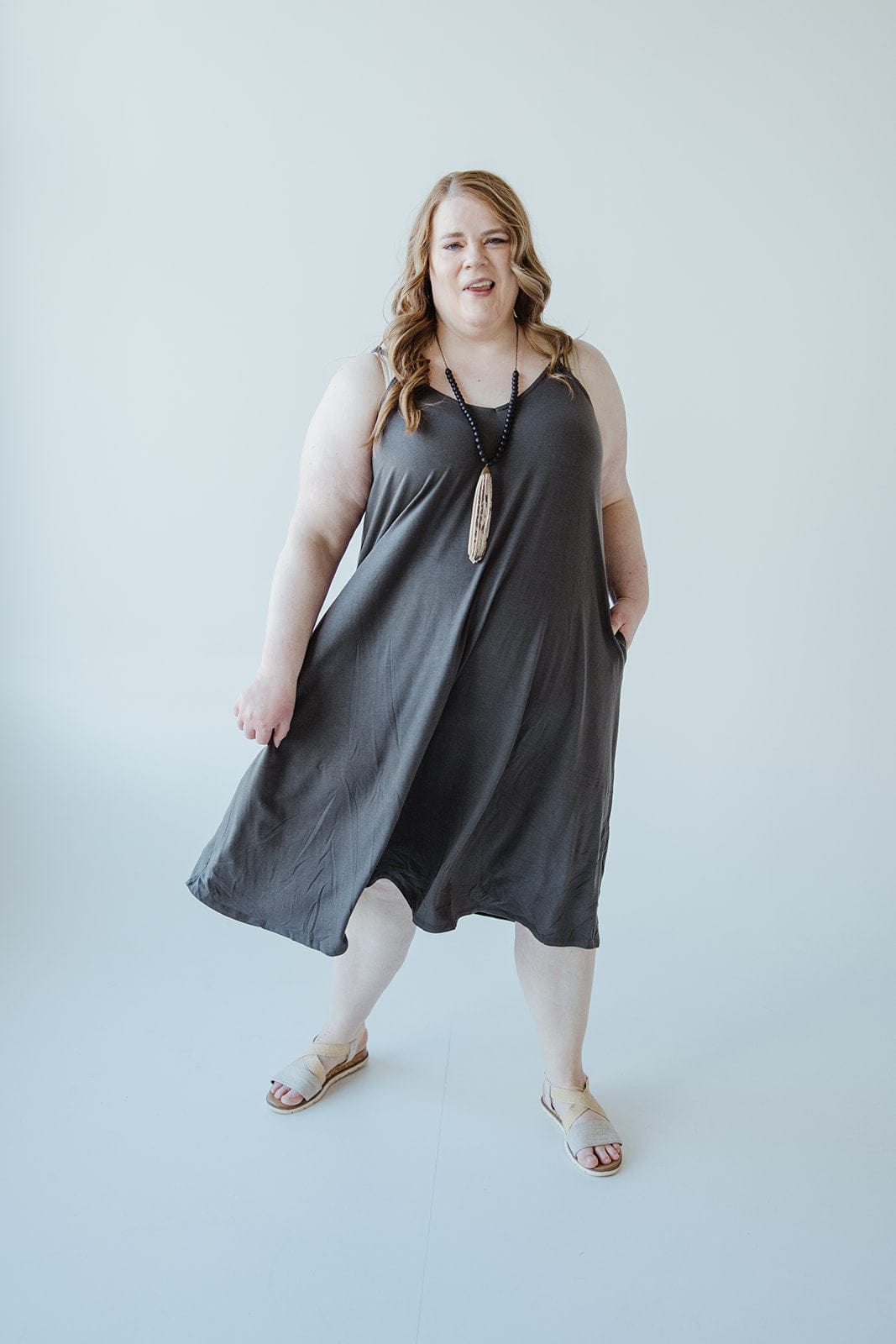 A person with long hair stands against a plain background, wearing a charcoal-hued cami dress paired with sandals.