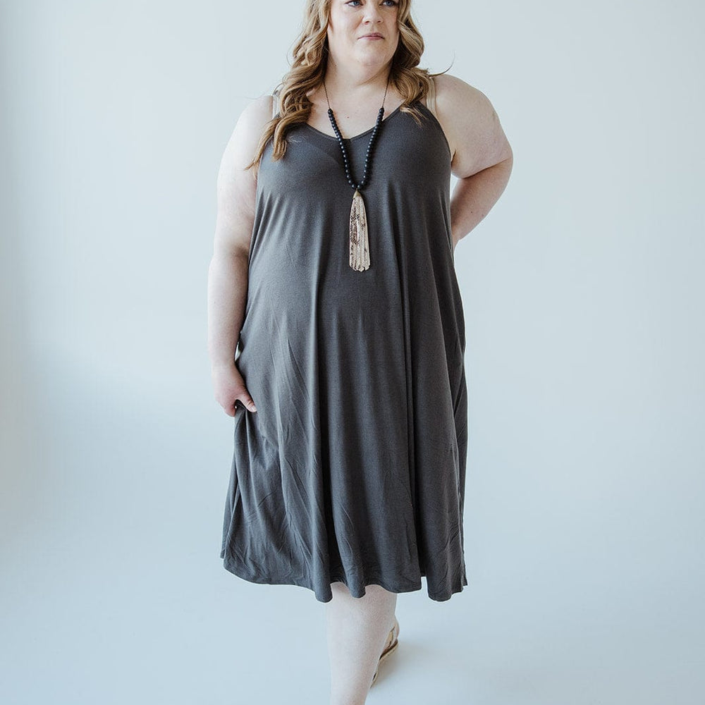 A woman in a charcoal-hued cami dress stands against a plain, light-colored background. She is adorned with a long necklace and beige sandals, perfectly complementing the elegance of her knee-length A-line ensemble.