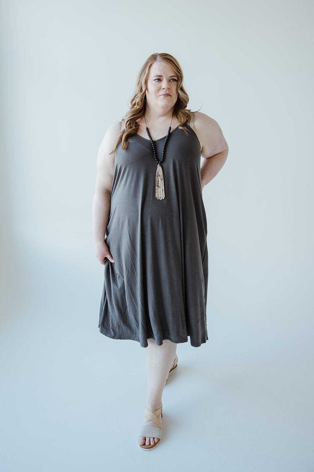 A woman in a charcoal-hued cami dress stands against a plain, light-colored background. She is adorned with a long necklace and beige sandals, perfectly complementing the elegance of her knee-length A-line ensemble.