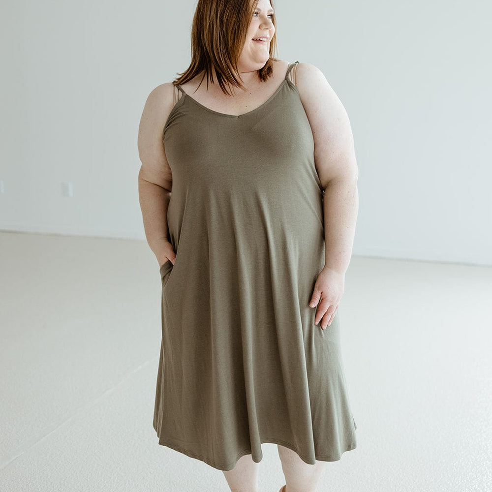 A person stands in a minimalist room wearing the Zenana Knee Length A-line Cami Dress in Mountain Olive with adjustable straps and brown sandals, looking to the side.