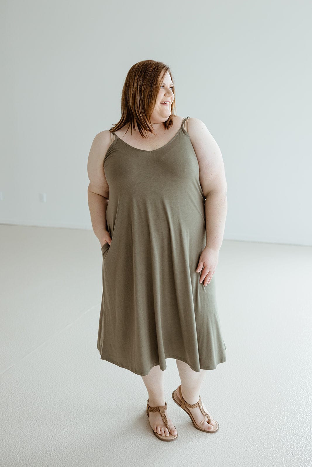 A person stands in a minimalist room wearing the Zenana Knee Length A-line Cami Dress in Mountain Olive with adjustable straps and brown sandals, looking to the side.