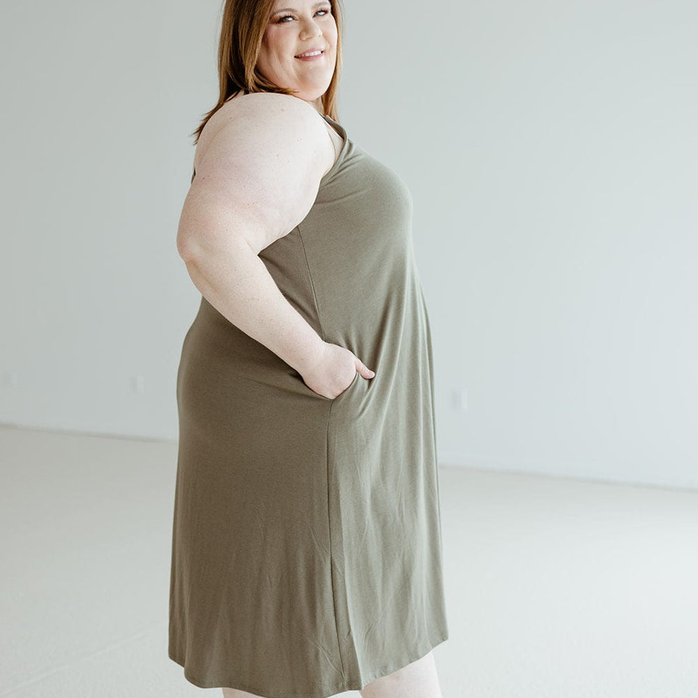 A woman wearing the Zenana Knee-Length A-Line Cami Dress in Mountain Olive paired with tan sandals is standing sideways with her hands in her pockets, looking at the camera and smiling.