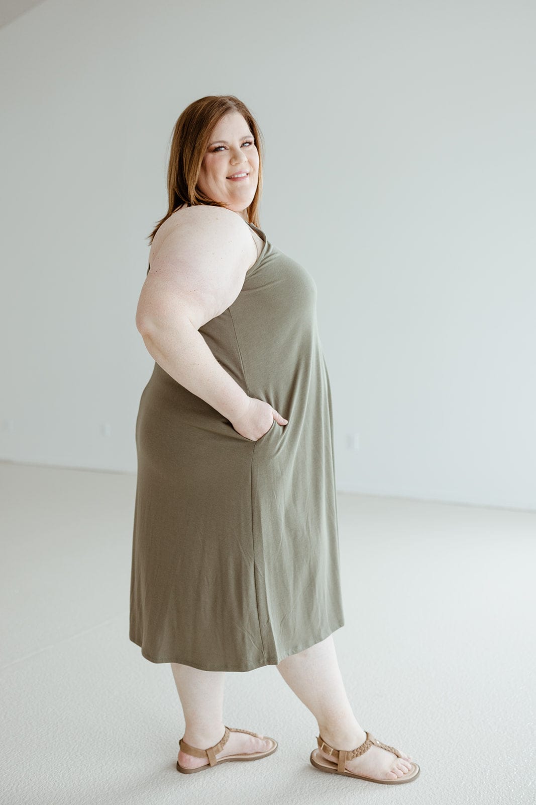 A woman wearing the Zenana Knee-Length A-Line Cami Dress in Mountain Olive paired with tan sandals is standing sideways with her hands in her pockets, looking at the camera and smiling.