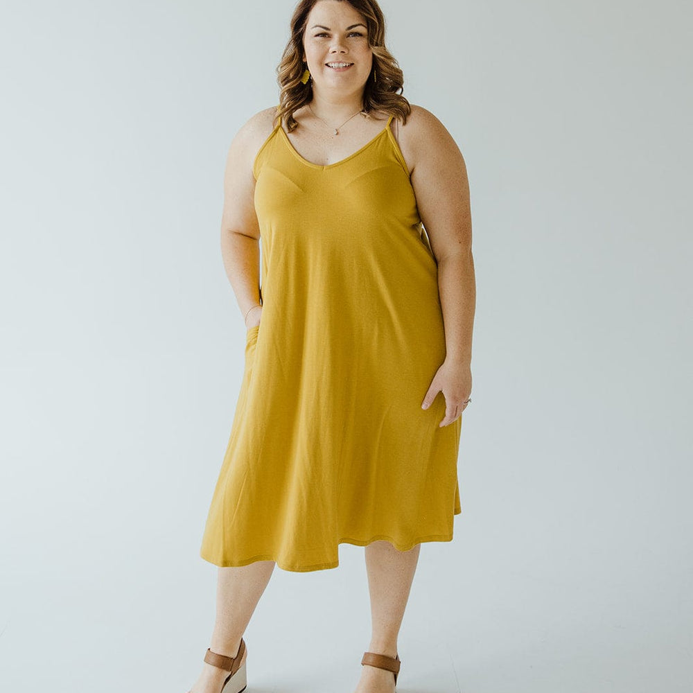 A person in a golden olive A-line cami dress with adjustable straps and sandals stands against a plain white background.