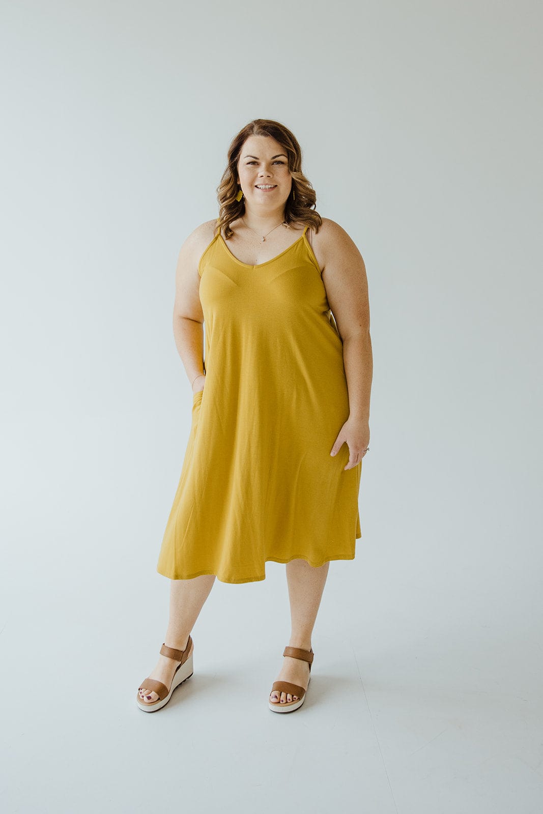 A person in a golden olive A-line cami dress with adjustable straps and sandals stands against a plain white background.