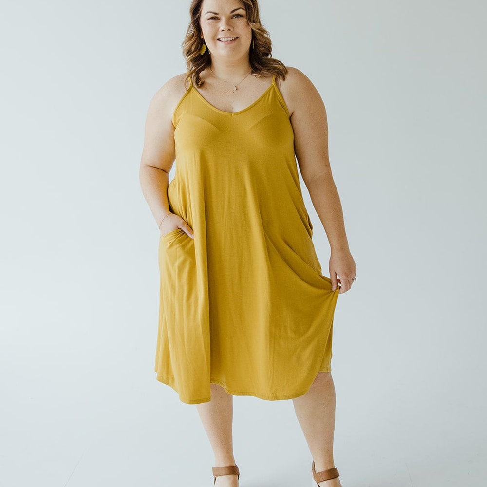 A person wearing an A-line cami dress in a vibrant golden olive hue stands in a bright space, smiling and posing with one hand casually tucked in the pocket, showcasing the chic charm of its adjustable straps.