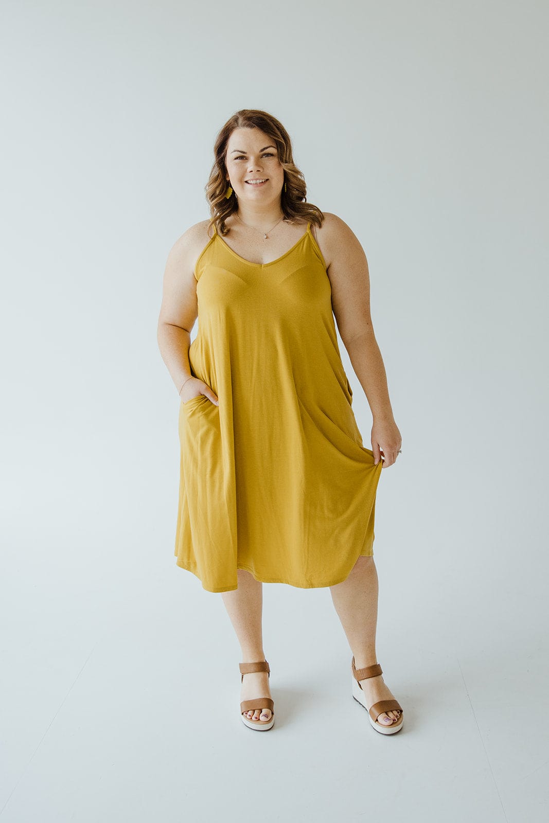 A person wearing an A-line cami dress in a vibrant golden olive hue stands in a bright space, smiling and posing with one hand casually tucked in the pocket, showcasing the chic charm of its adjustable straps.