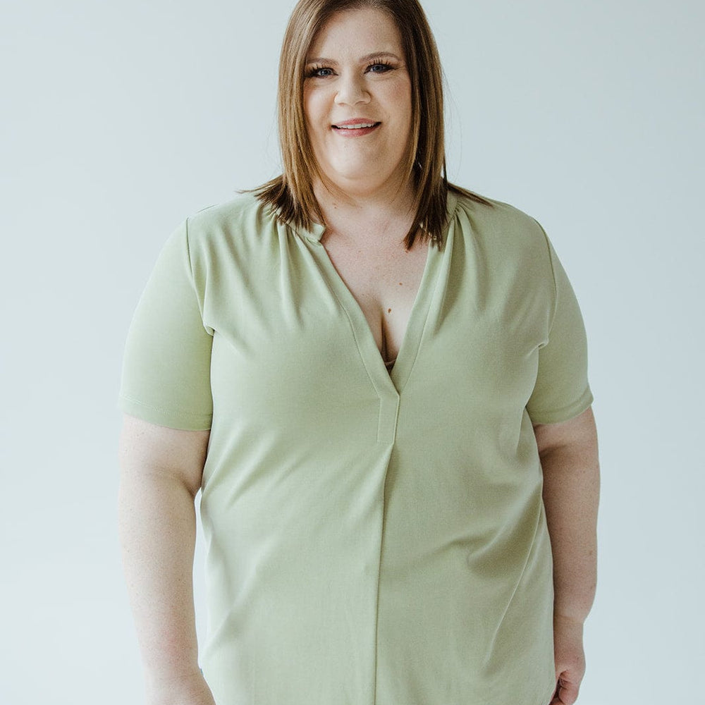 A person with shoulder-length brown hair wearing a light green KNIT BLOUSE WITH MANDARIN COLLAR by Love Marlow and jeans stands against a plain white background, smiling at the camera.