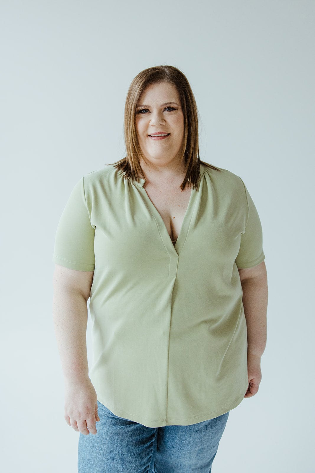 A person with shoulder-length brown hair wearing a light green KNIT BLOUSE WITH MANDARIN COLLAR by Love Marlow and jeans stands against a plain white background, smiling at the camera.