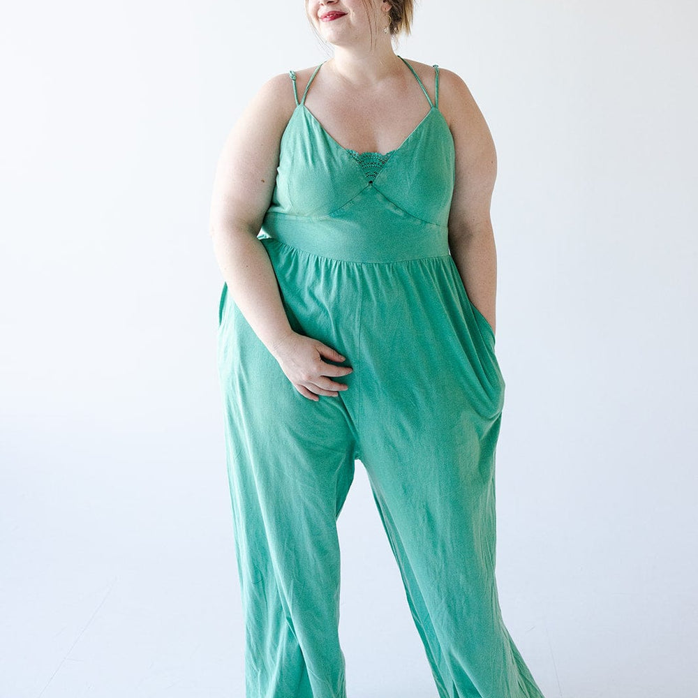 A woman stands against a plain white background, wearing a loose, green KNIT JUMPSUIT WITH PEEK-A-BOO LACE by Love Marlow and black sandals. The summer vibes are evident as she has her hands in the pockets of the jumpsuit and gazes to the side with a slight smile.