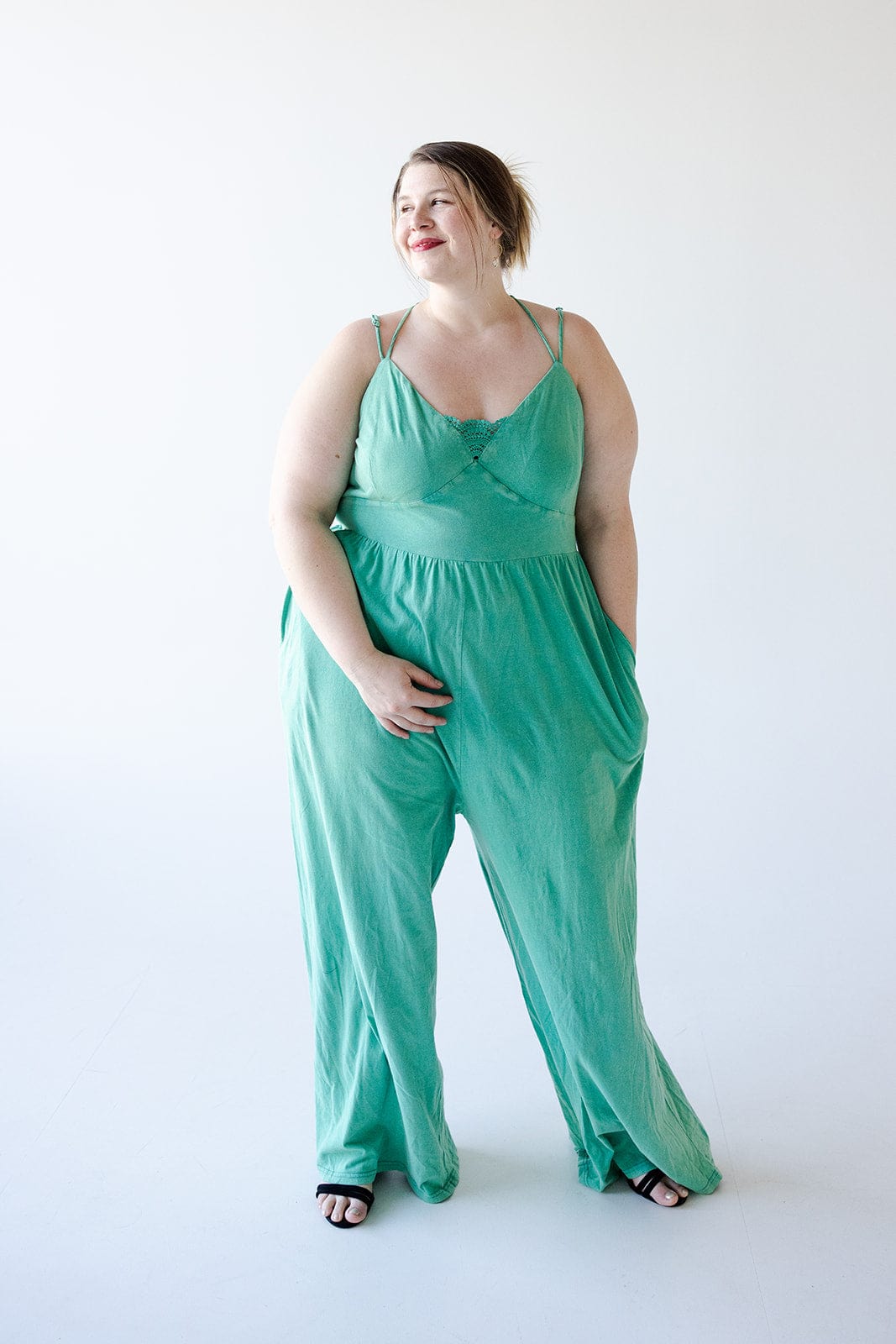 A woman stands against a plain white background, wearing a loose, green KNIT JUMPSUIT WITH PEEK-A-BOO LACE by Love Marlow and black sandals. The summer vibes are evident as she has her hands in the pockets of the jumpsuit and gazes to the side with a slight smile.