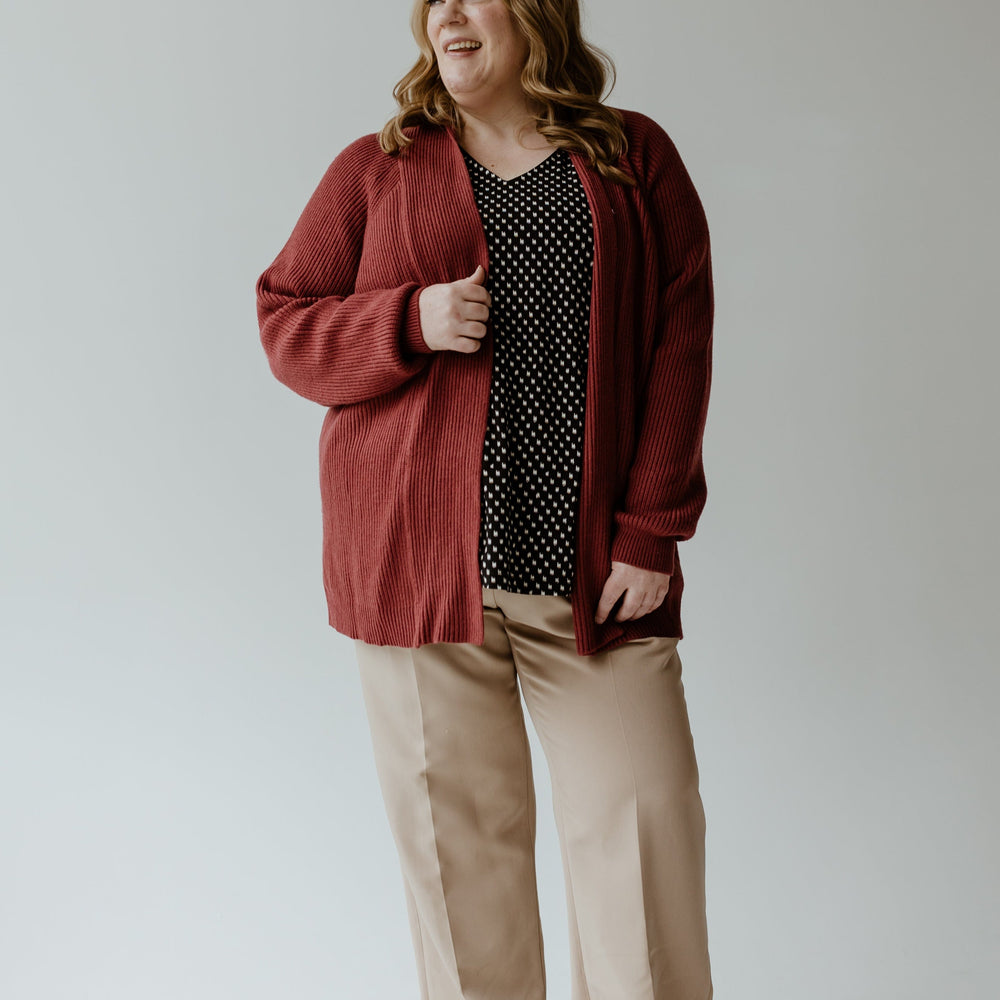 A person stands smiling in a Yesta KNIT OPEN CARDIGAN IN CRANBERRY, paired with a black polka dot blouse, beige pants, and dark shoes against a plain background.