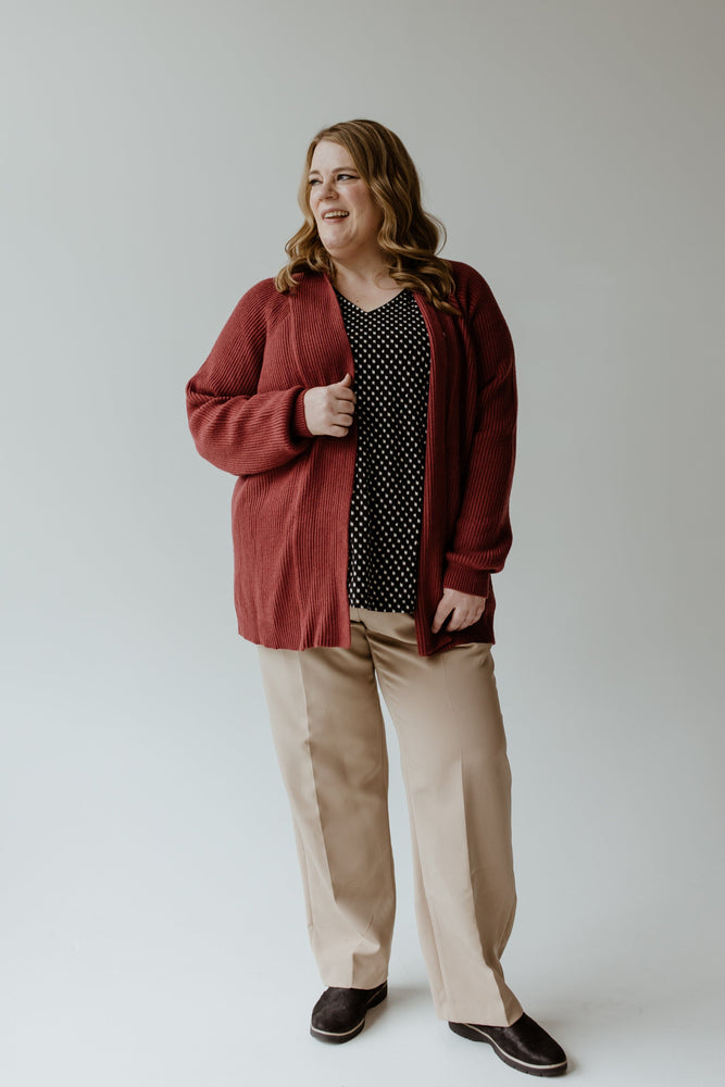 A person stands smiling in a Yesta KNIT OPEN CARDIGAN IN CRANBERRY, paired with a black polka dot blouse, beige pants, and dark shoes against a plain background.