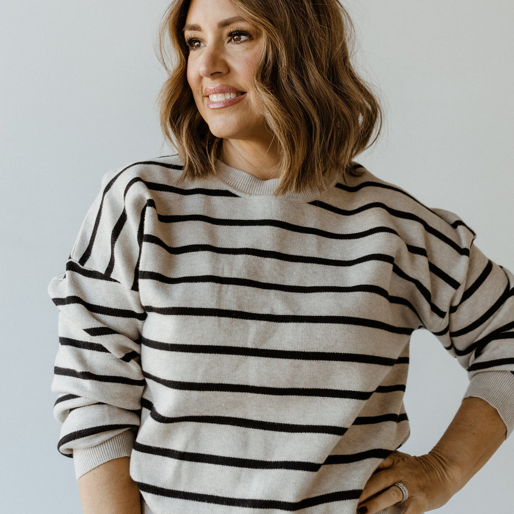 A woman with shoulder-length hair and a smile dons a knit striped sweater with dolman blouson sleeves, standing confidently with one hand on her hip against a plain background.