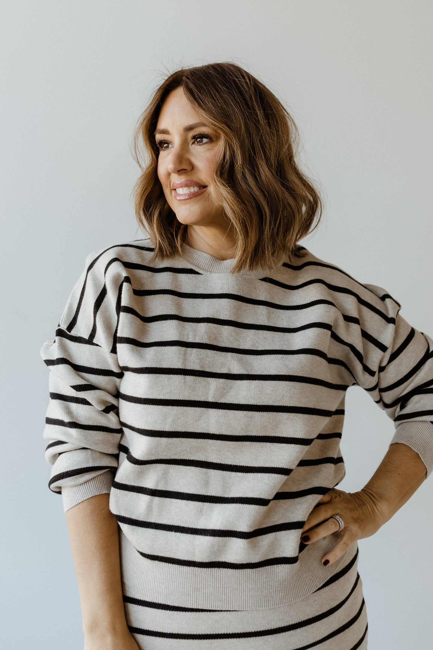 A woman with shoulder-length hair and a smile dons a knit striped sweater with dolman blouson sleeves, standing confidently with one hand on her hip against a plain background.