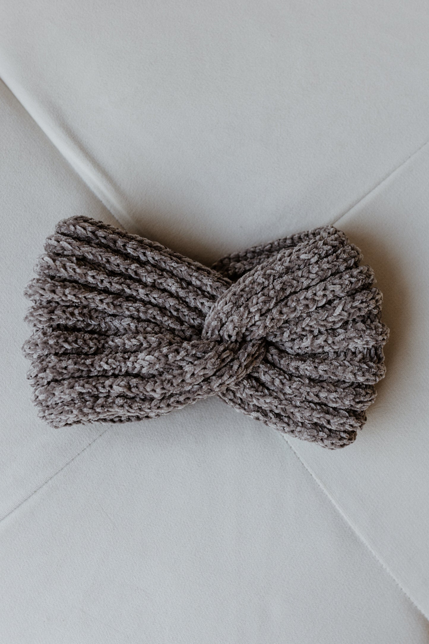 A knitted chenille headband with a twisted design rests elegantly on a white surface, perfect for adding warmth and style to any winter outfit.