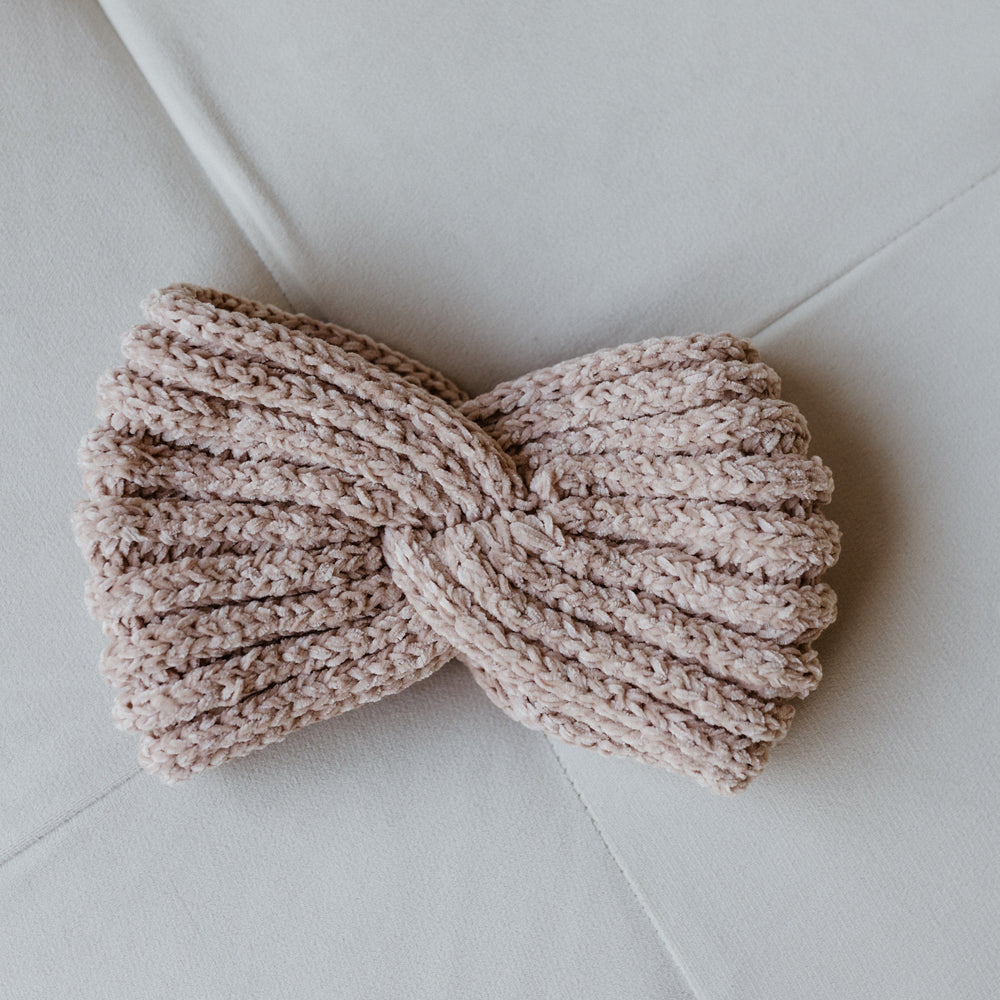 A stylish knitted chenille headband with a twisted design rests on a light gray surface, offering winter warmth and charm.