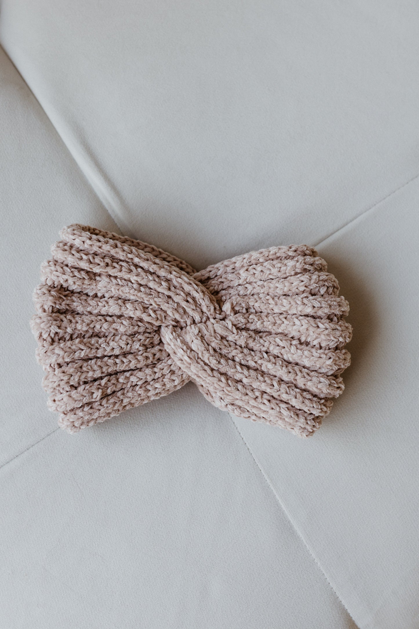 A stylish knitted chenille headband with a twisted design rests on a light gray surface, offering winter warmth and charm.