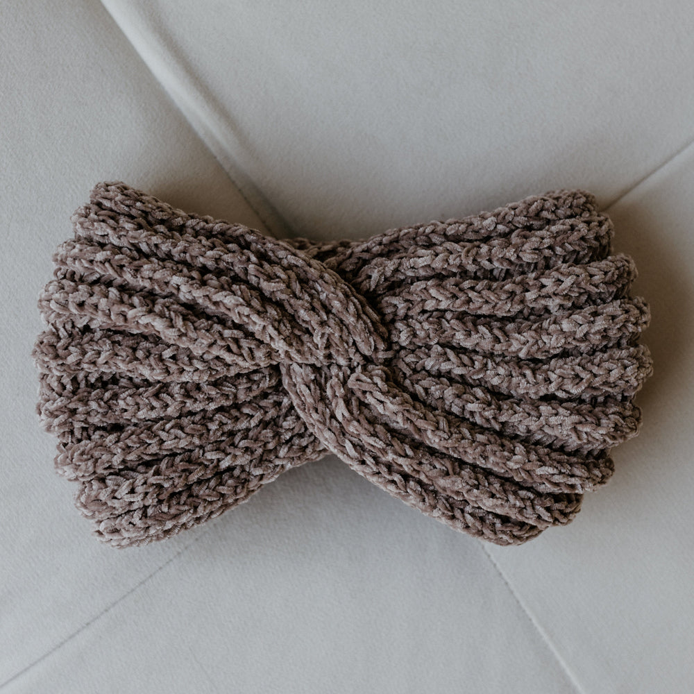 A super soft, knitted chenille headband featuring a twisted knot design lies on a light gray surface, perfect for those cozy winter days.