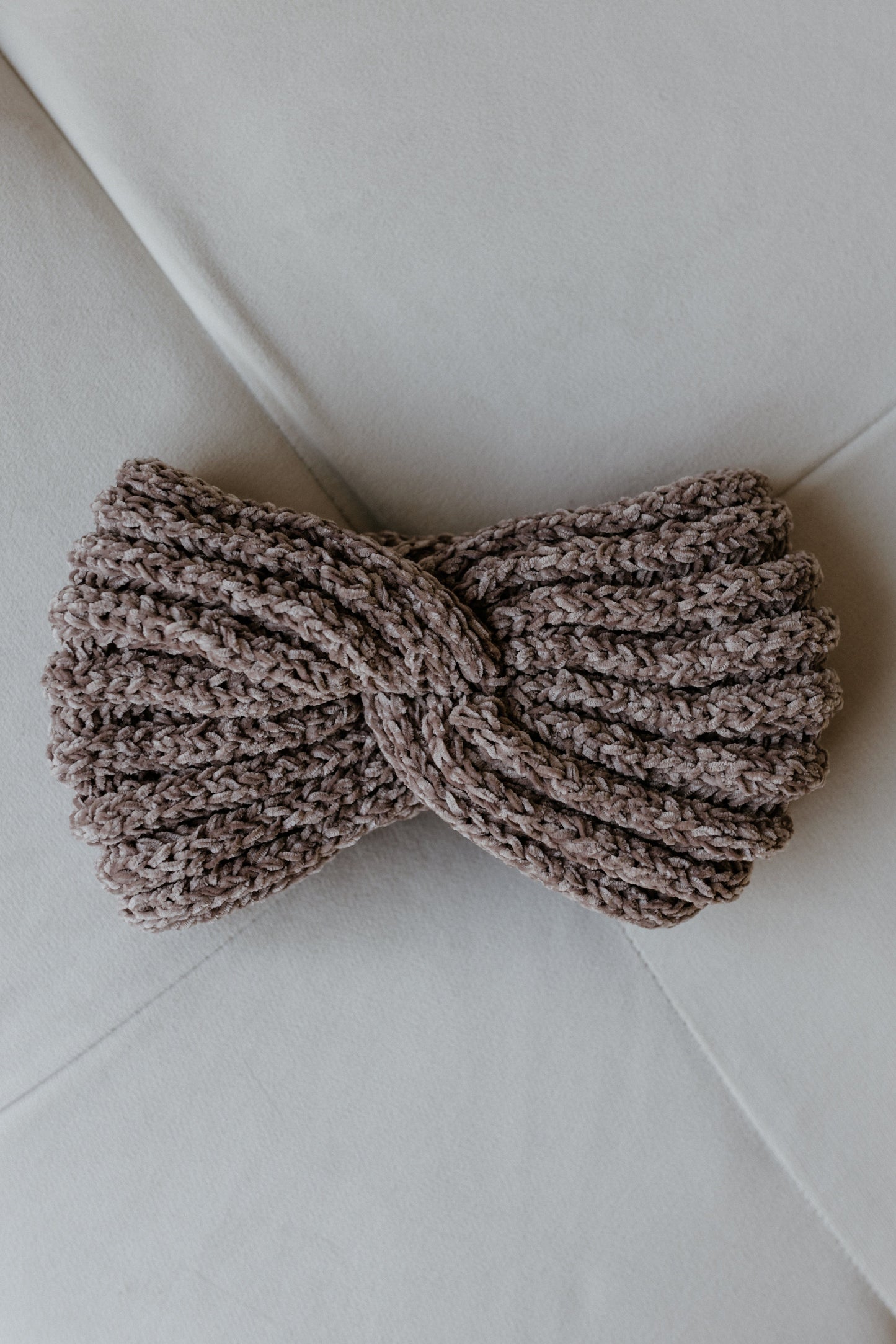 A super soft, knitted chenille headband featuring a twisted knot design lies on a light gray surface, perfect for those cozy winter days.