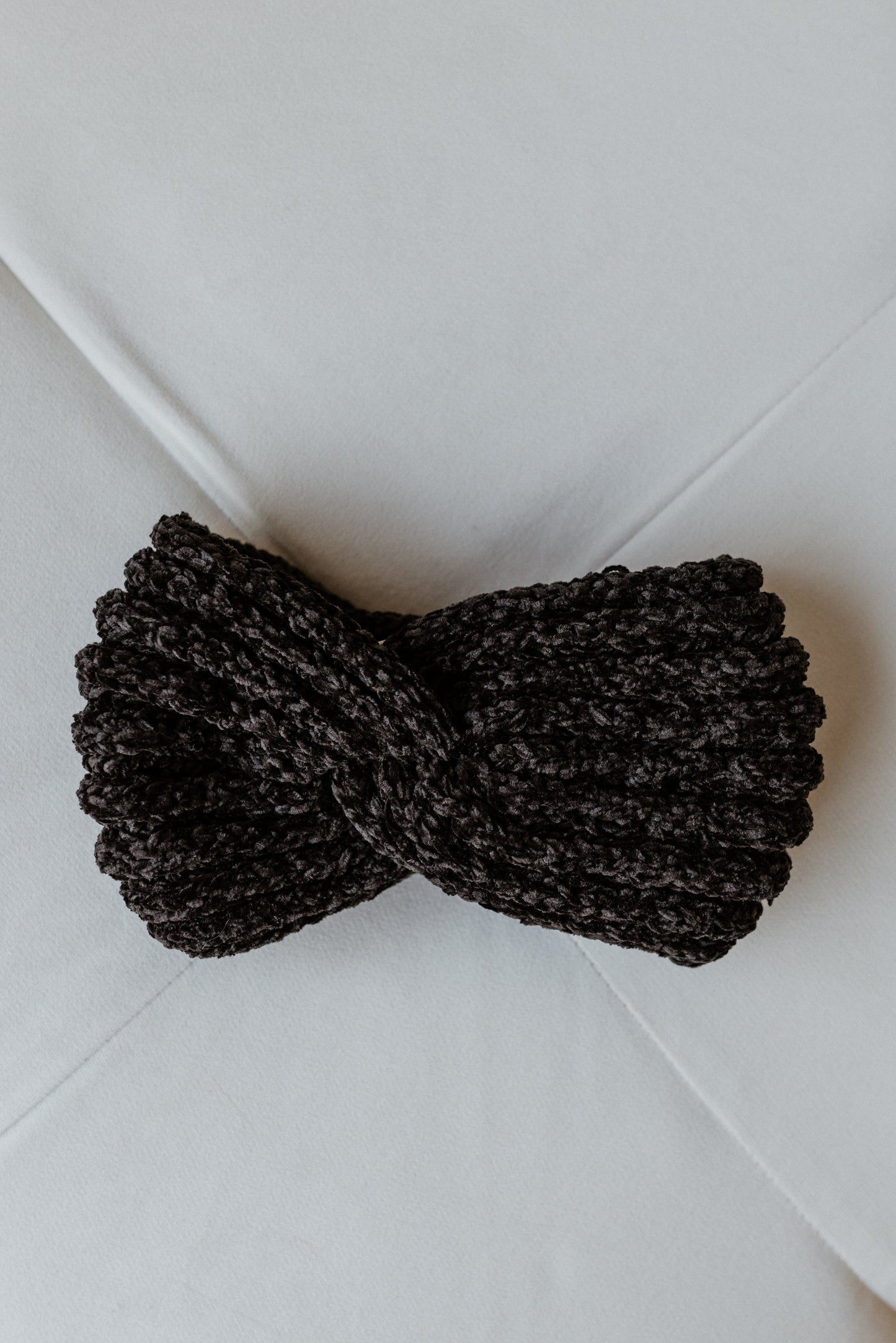 A black knitted chenille headband with a twisted design lies on a white surface, exuding an air of cozy elegance.