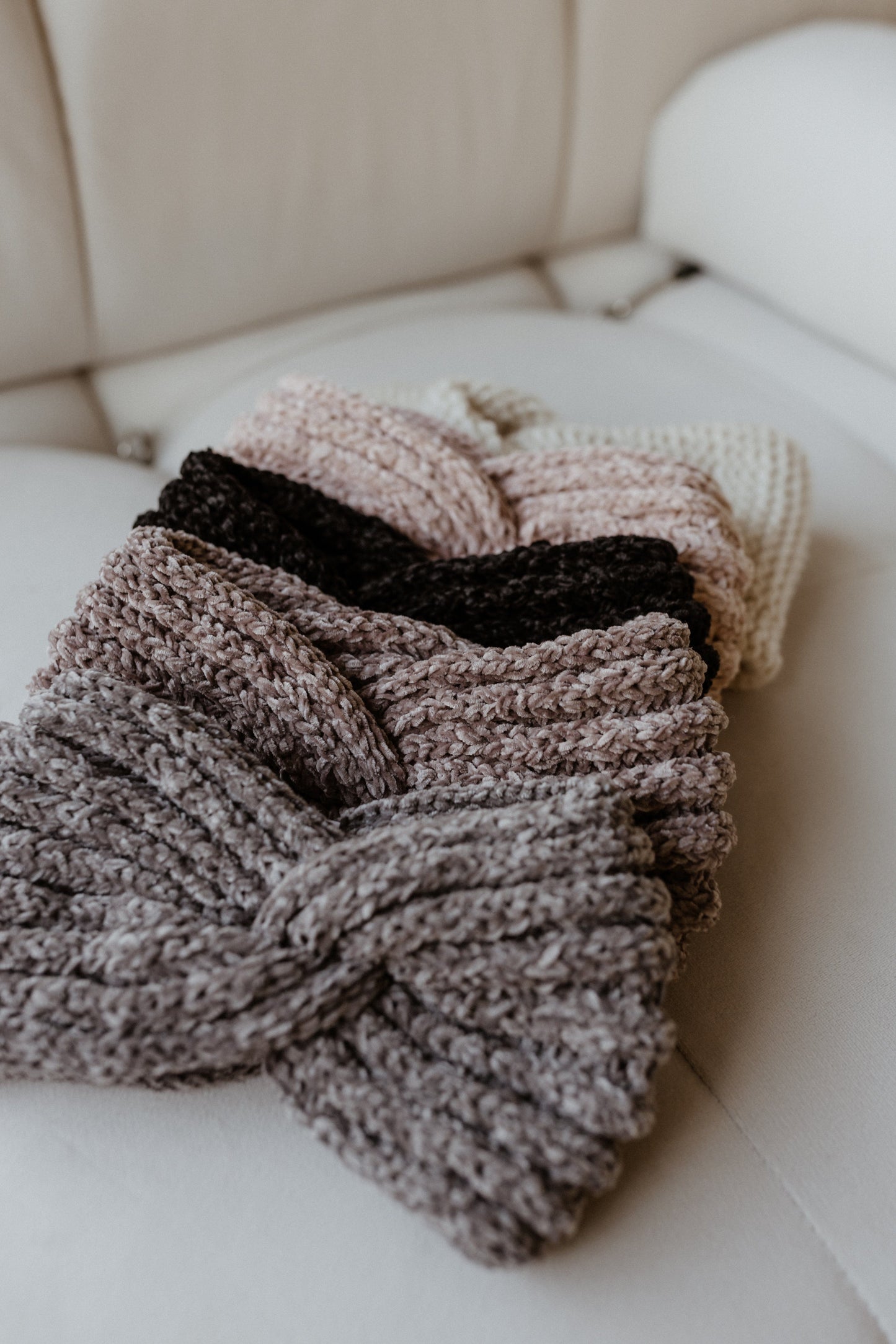 A stack of super-soft knitted chenille headbands in neutral tones is beautifully laid out on a pristine white couch.