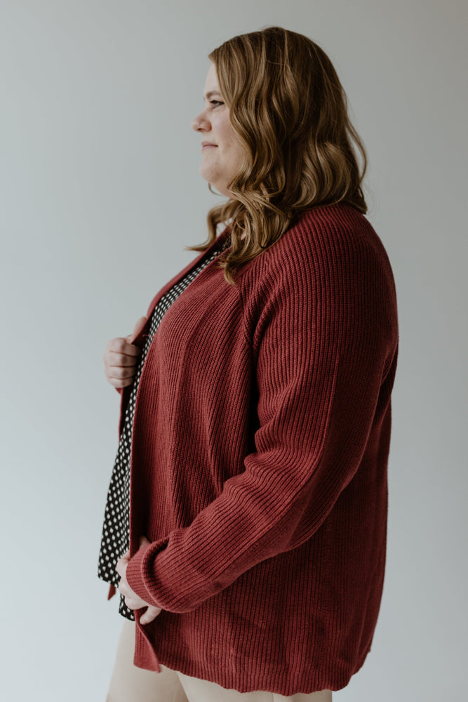 A woman with wavy brown hair is wearing the Yesta KNIT OPEN CARDIGAN IN CRANBERRY over a polka dot top, standing sideways against a plain background.