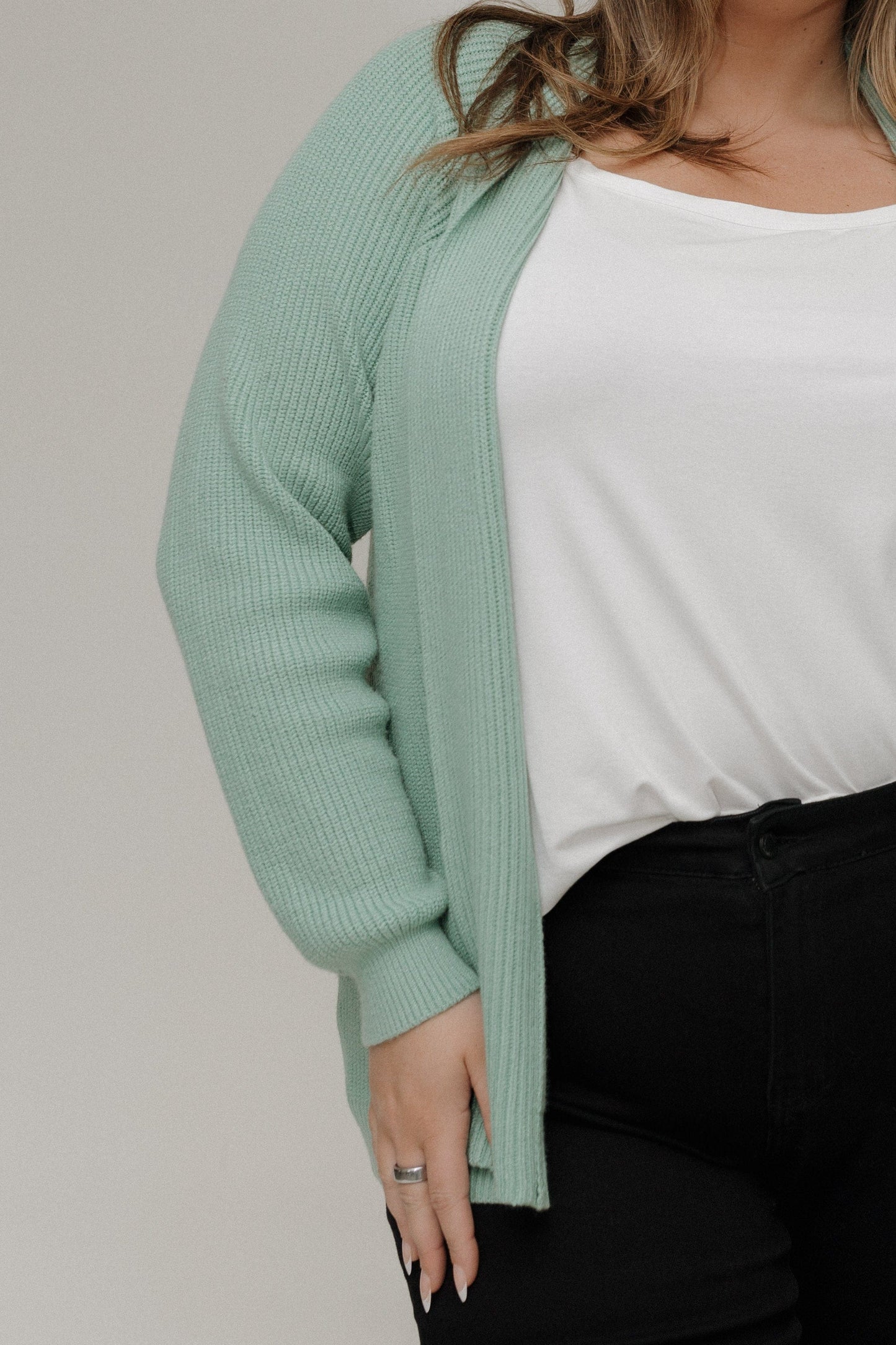 Person wearing a light green cardigan over a white shirt and black pants on a plain background