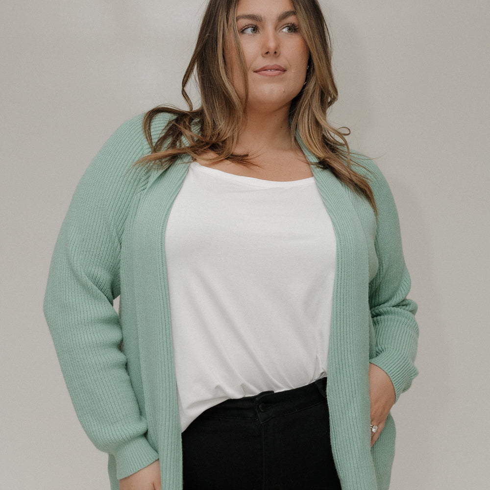 Woman wearing a light green cardigan over a white top and black pants against a plain background