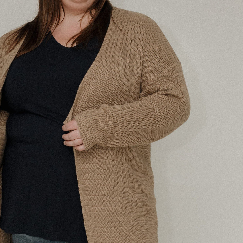 Woman wearing a beige cardigan over a black top and blue jeans against a plain background