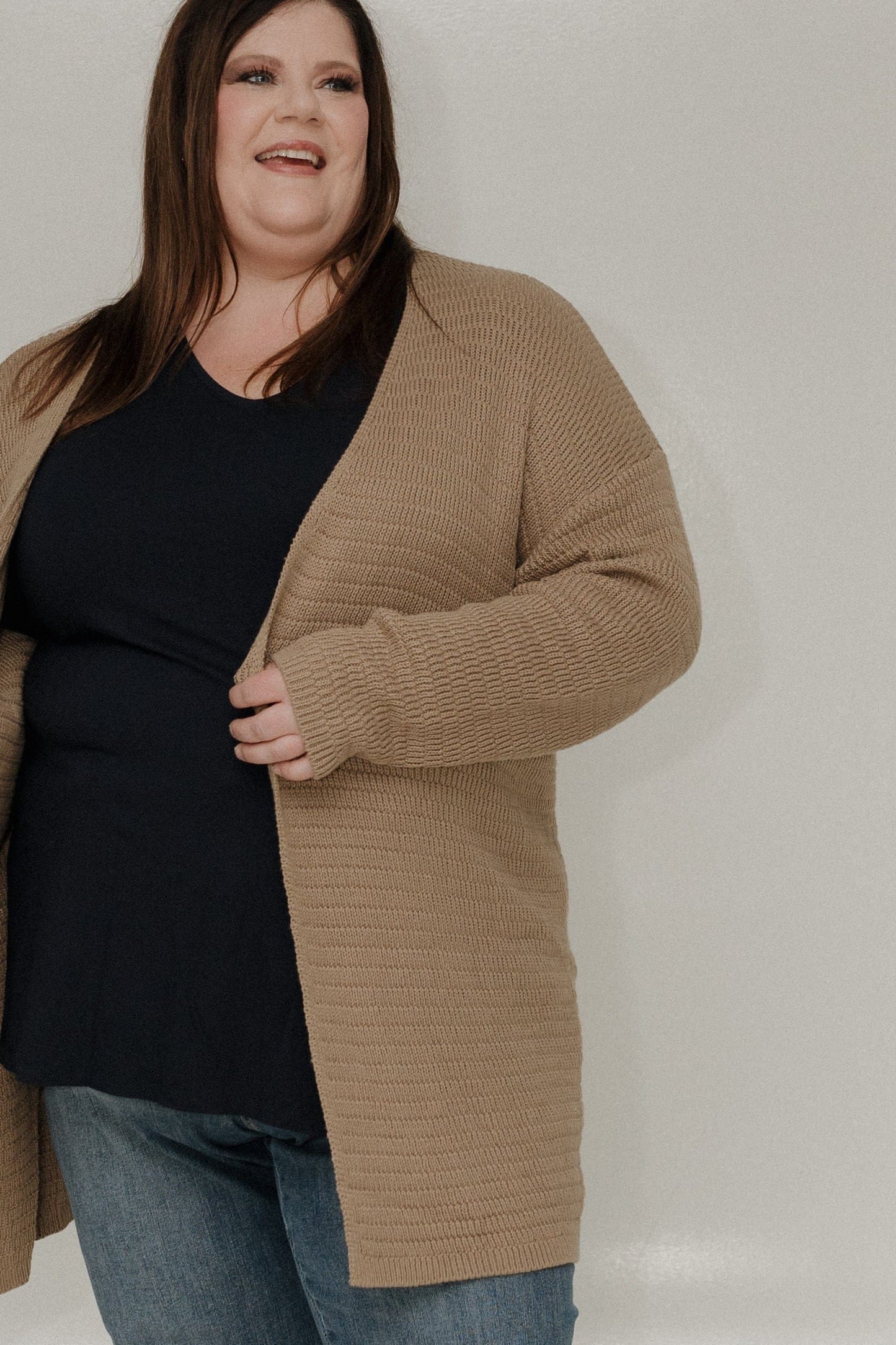 Woman wearing a beige cardigan over a black top and blue jeans against a plain background