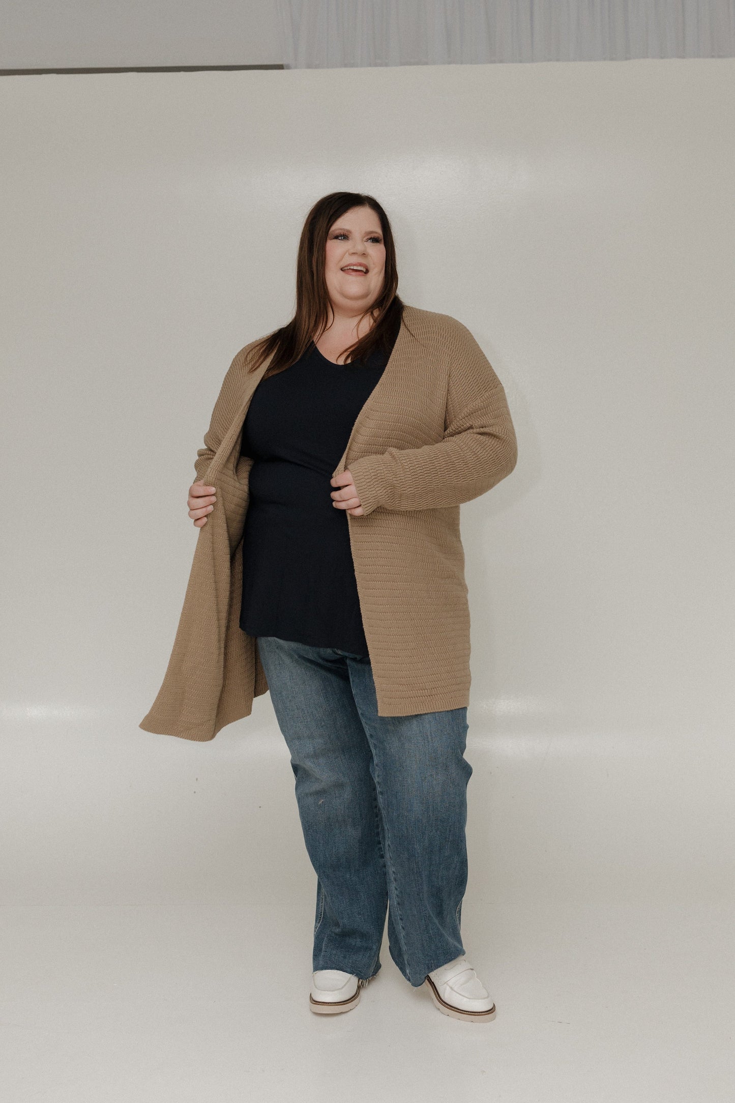 Person wearing a beige coat, black top, blue jeans, and white shoes on a plain background