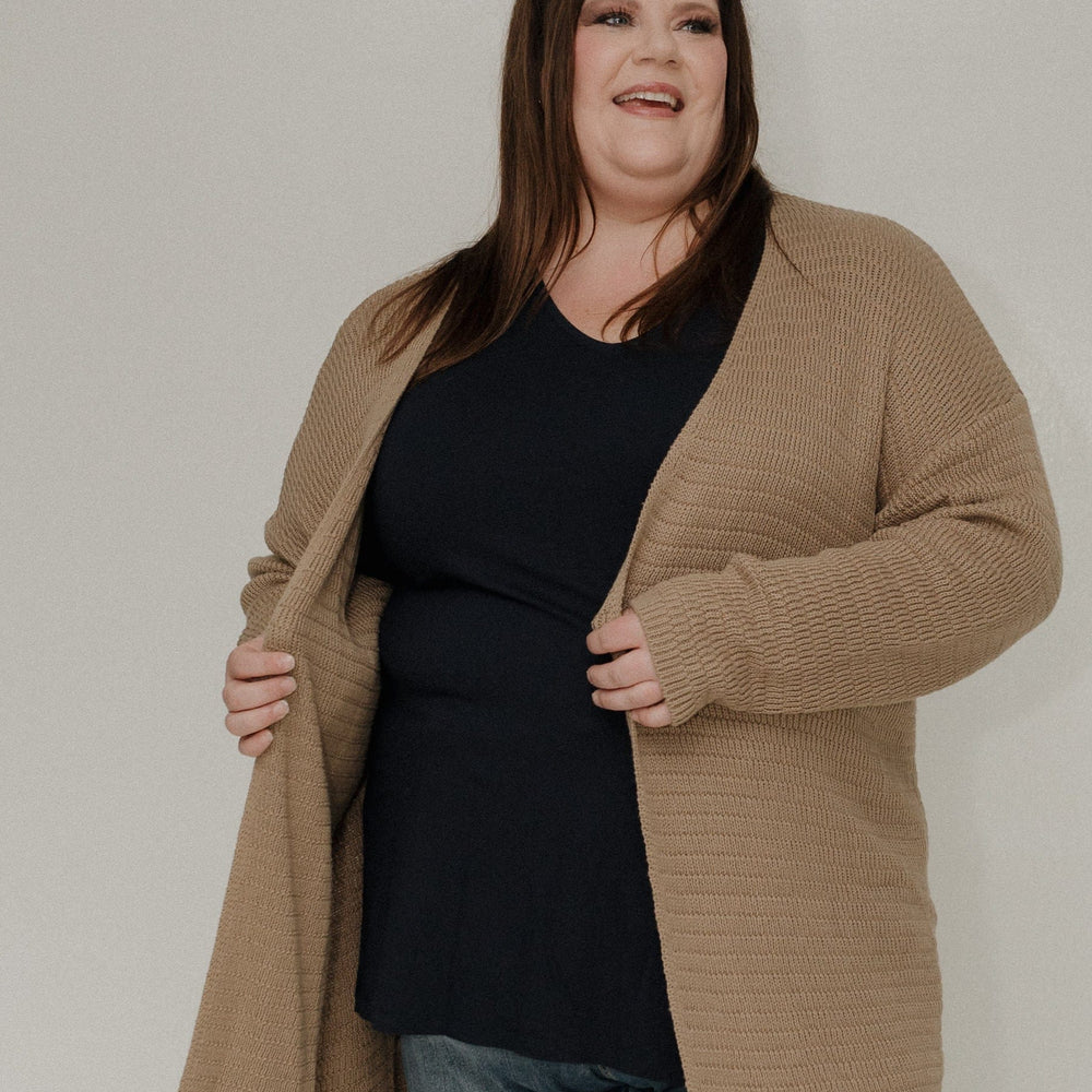 Person wearing a beige coat, black top, blue jeans, and white shoes on a plain background