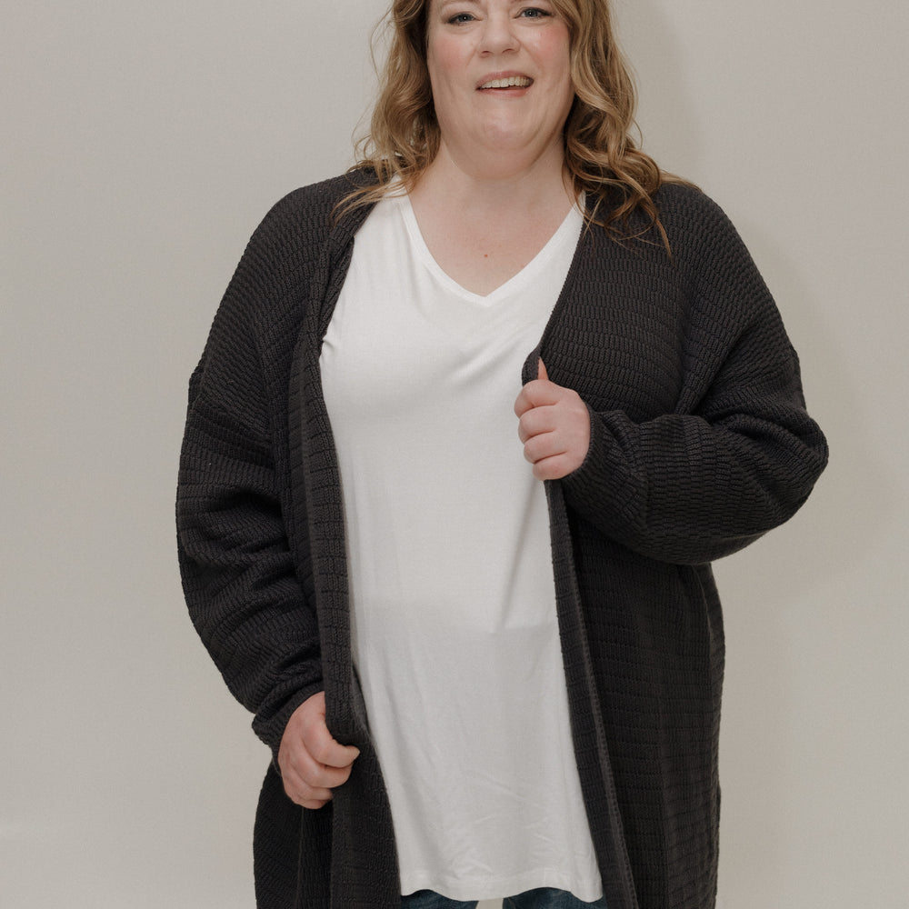 Person wearing a black cardigan over a white shirt and blue jeans on a plain background